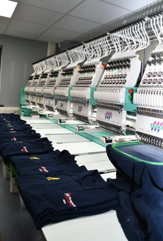 Multi-head embroidery machine next to a stack of navy polo shirts with embroidered logos