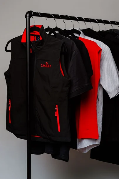 Gilet i zi me zinxhirë të kuq, polo dhe këmisha uniforme të varura në një stendë rrobash