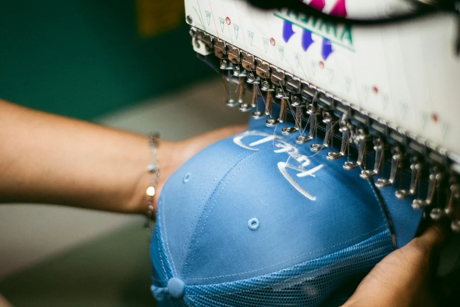 Blue cap held under a multi-head embroidery machine while a logo is stitched onto the front